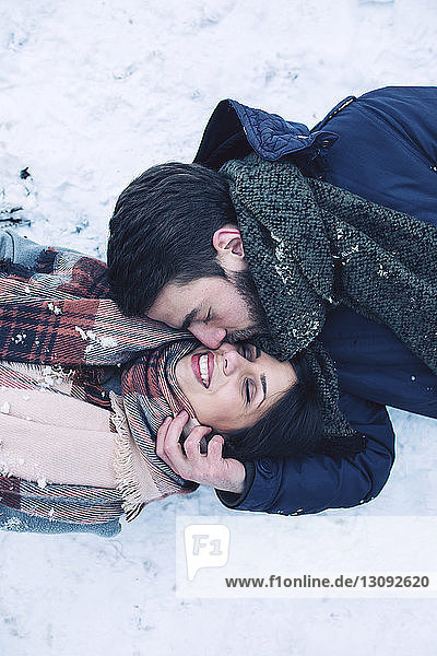 High angle view of man kissing girlfriend while lying on snow covered field