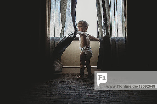 Rear view of shirtless baby boy playing with curtains while standing by window at home