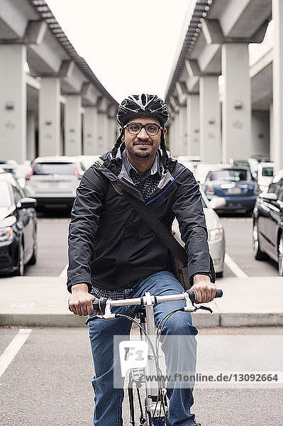 Porträt eines lächelnden  auf dem Fahrrad sitzenden Geschäftsmannes mit Brücken und Parkplatz im Hintergrund