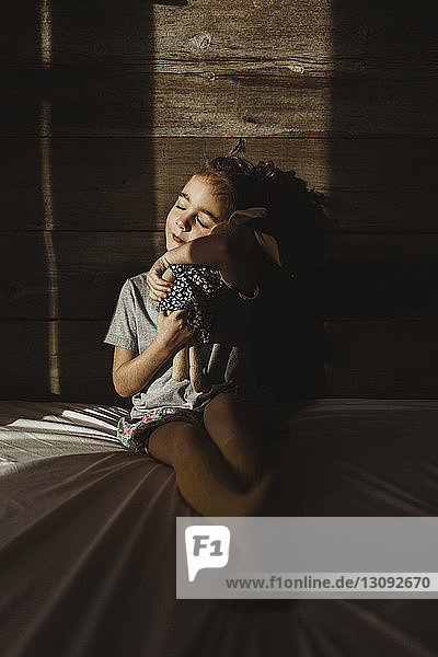 Mädchen mit geschlossenen Augen umarmt Stoffspielzeug  während sie in der Dunkelkammer auf dem Bett gegen Holz sitzt