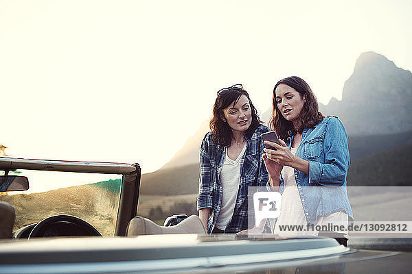 Frau zeigt Freundin Mobiltelefon im Cabriolet bei Sonnenuntergang