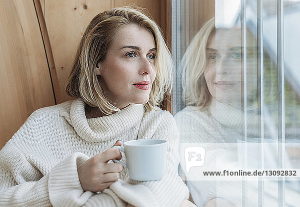 Frau hält Kaffeetasse  während sie zu Hause durchs Fenster schaut