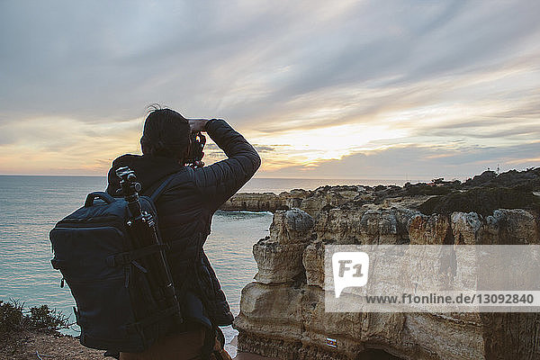 Rear view of man photographing sea while standing on rock formation against cloudy sky