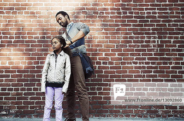 Father tying daughter's hair while standing against brick wall