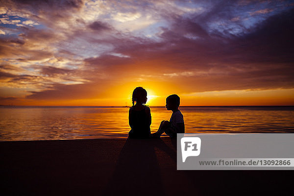 Silhouette siblings sitting by beach during sunset