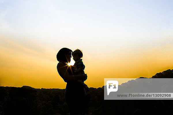 Silhouette einer Mutter  die einen Sohn trägt  während sie bei Sonnenuntergang im Park gegen den Himmel steht