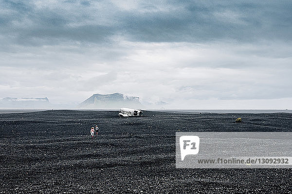 Mitteldistanzansicht der Geschwister  die auf das Flugzeugwrack auf schwarzem Sand am Strand vor bewölktem Himmel zulaufen