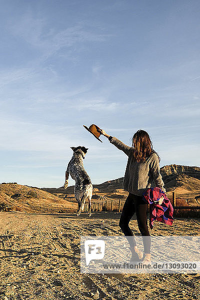 Full length of young woman playing with dog at desert against sky