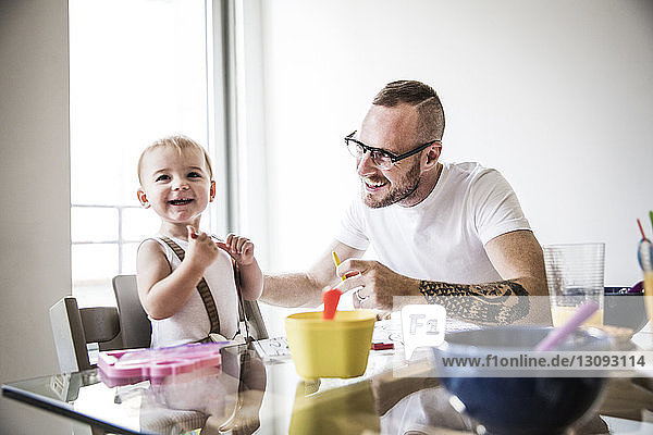 Happy father feeding daughter at breakfast table against wall
