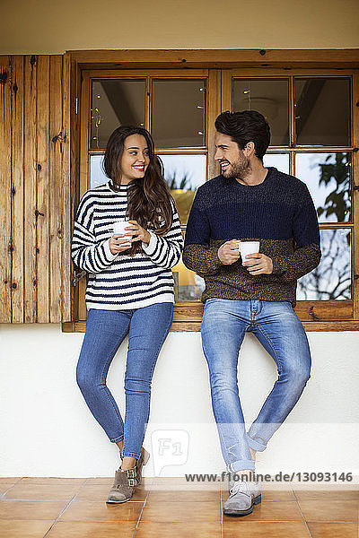 Lächelndes Paar trinkt Kaffee  während es am Fenster auf der Veranda steht