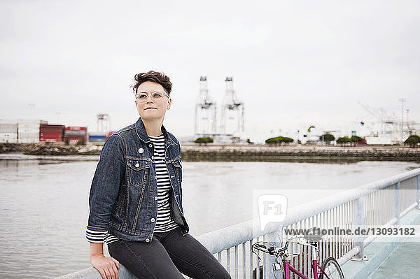 Woman with bicycle looking away while sitting on railing against sea