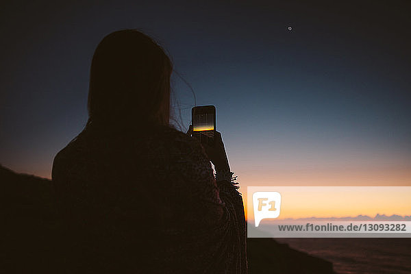 Rückansicht einer Frau  die den Sonnenuntergang mit einem Smartphone fotografiert
