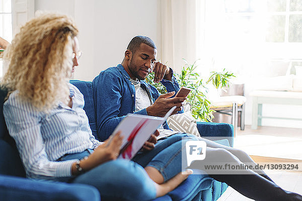 Frau liest Zeitschrift  während der Mann zu Hause auf dem Sofa ein Smartphone benutzt
