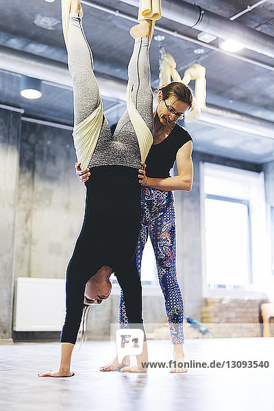 Yogalehrerin assistiert Frau, die in der Hängematte hängt, während sie im Fitnessstudio trainiert