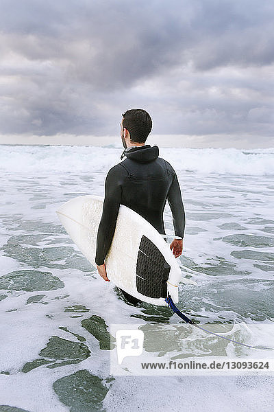 Rear view of man carrying surfboard while walking in sea against cloudy sky