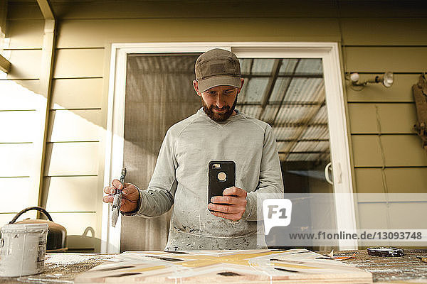 Selbstbewusster Künstler fotografiert Holzkunst mit dem Handy an der Werkbank