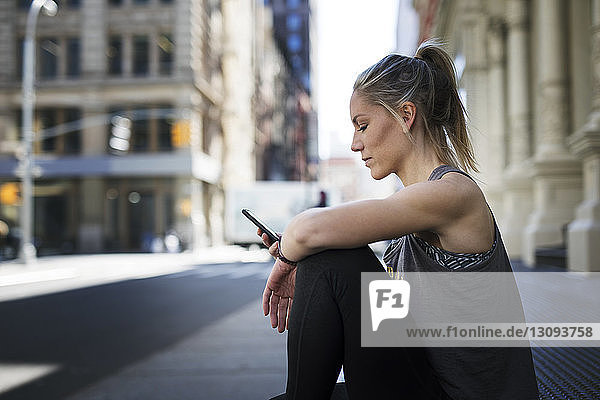 Sportler benutzt Mobiltelefon  während er in der Stadt auf dem Bürgersteig sitzt