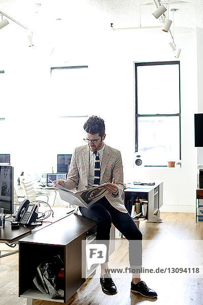 Full length of businessman reading newspaper while sitting on desk in office