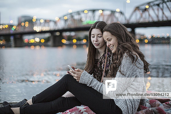 Lächelnde Freundinnen  die ein Smartphone benutzen  während sie auf dem Pier gegen die Burlington Northern Railroad Bridge sitzen 9 6