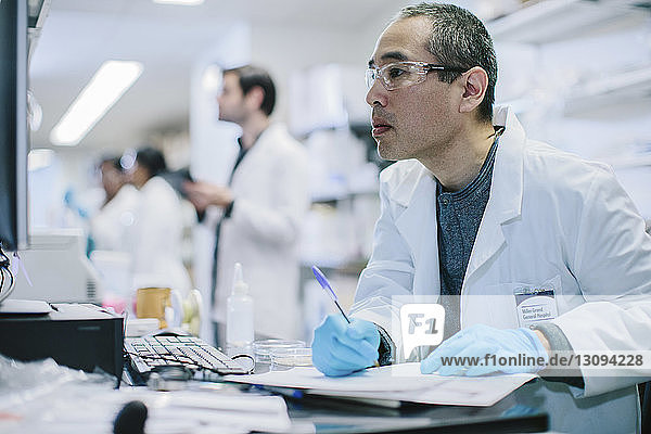 Male doctor writing at desk while coworkers working in background