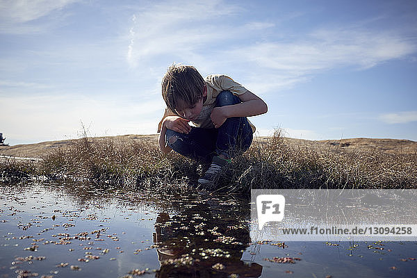 Boy looking into lake while crouching on grassy field against sky