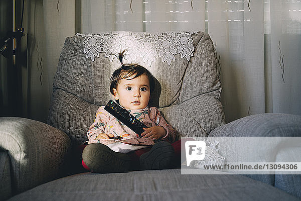 Cute baby girl with remote control sitting on armchair at home
