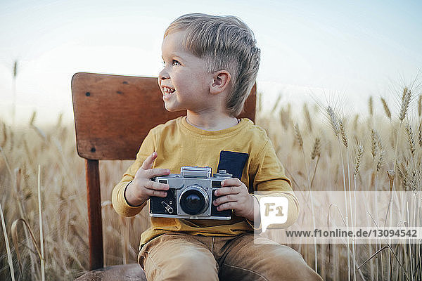 Cheerful boy with camera looking away while sitting on chair amidst wheat field