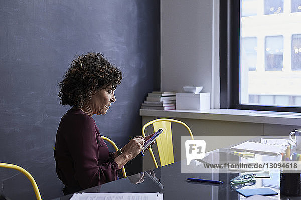 Ältere Geschäftsfrau benutzt Mobiltelefon  während sie im Kreativbüro am Konferenztisch sitzt