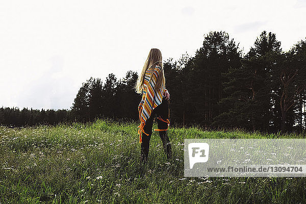 Rückansicht einer Frau in Poncho  die auf einem Grasfeld vor klarem Himmel steht