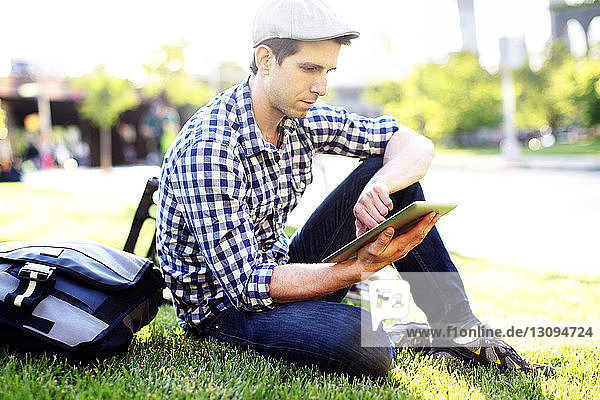 Mann benutzt digitales Tablett  während er an einem sonnigen Tag auf einem Grasfeld sitzt