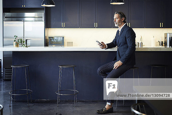 Ein Geschäftsmann in voller Länge schaut weg  während er ein Smartphone an der Küchentheke im Kreativbüro hält