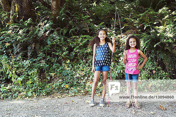 Portrait of sisters with fishing rods standing footpath against trees