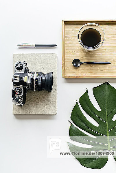 Draufsicht auf Kaffee in Holztablett mit Kamera und Büromaterial durch Monsterblatt auf weißem Hintergrund