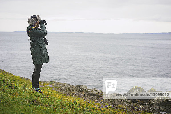 Woman looking through binoculars while standing on field by sea against sky