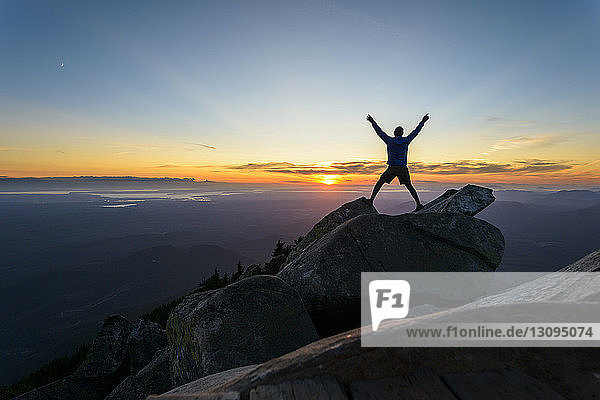 Scherenschnitt eines Mannes mit erhobenen Armen  der bei Sonnenuntergang im Mount Pilchuck State Park auf Felsen gegen den Himmel steht