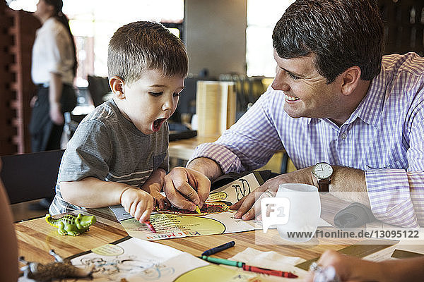 Happy father looking at shocked boy while coloring menu in restaurant