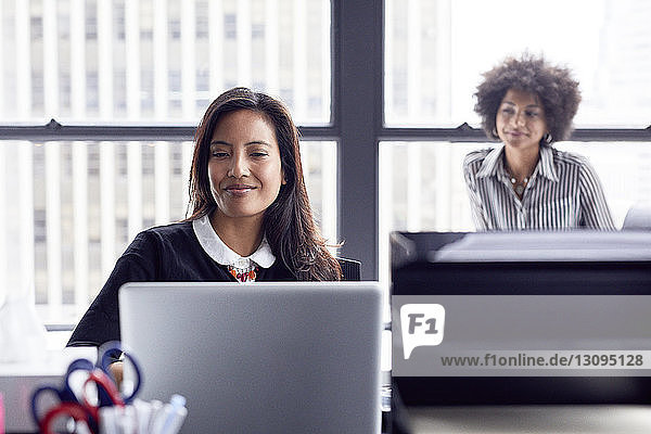 Geschäftsfrau benutzt Laptop-Computer  während sie im Büro mit einer Kollegin im Hintergrund gegen das Fenster arbeitet