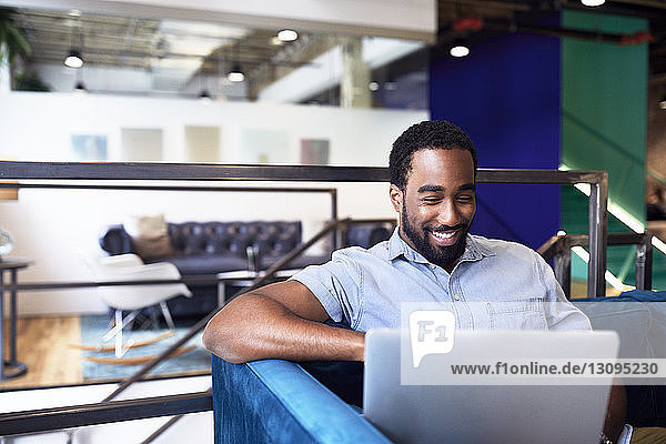 Lächelnder junger Geschäftsmann mit Laptop  während er im Büro auf dem Sofa sitzt