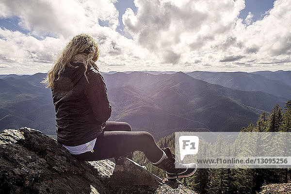 Woman sitting on rock at mountain cliff against cloudy sky