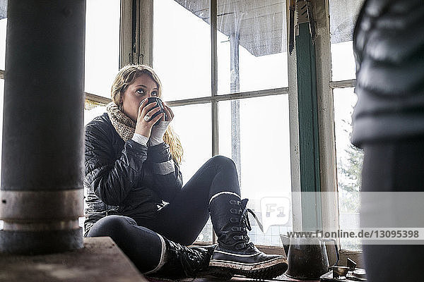 Frau sitzt am Fenster und trinkt Kaffee in Hütte