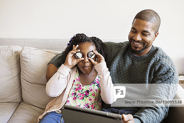 Mädchen spielt mit Keksen  während sie mit dem Vater am Tablet-Computer auf dem Sofa sitzt