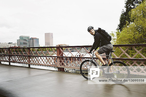 Seitenansicht eines männlichen Pendlers auf einer Brücke über einen Fluss mit dem Fahrrad gegen den Himmel