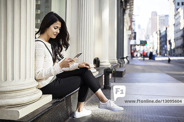 Young woman using smart phone while sitting by columns on sidewalk