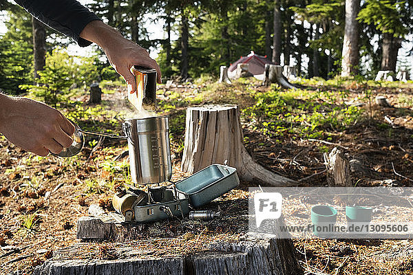 Beschnittenes Bild eines Mannes  der im Wald Lebensmittel in einen Behälter über einen Campingkocher schüttet
