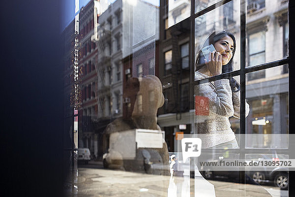 Young woman answering smart phone seen through glass wall
