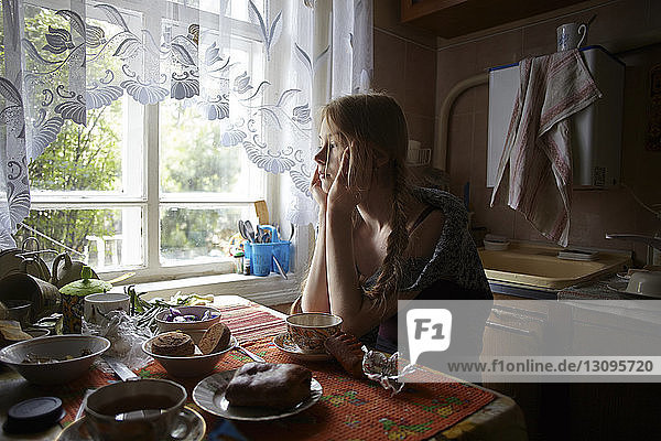 Thoughtful woman with hands on chin looking away while sitting by table at home