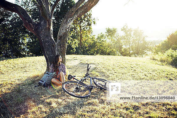 Junge Frau lehnt auf Baumstamm  während sie auf Grasfeld sitzt