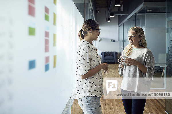 Lächelnde Geschäftsfrau trinkt Kaffee  während sie einem am Gang stehenden Kollegen zuhört