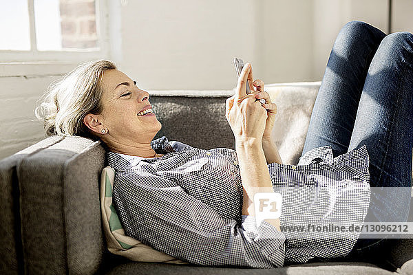 Happy woman using smart phone while lying on sofa at home