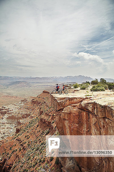Couple with bicycles on cliff against cloudy sky